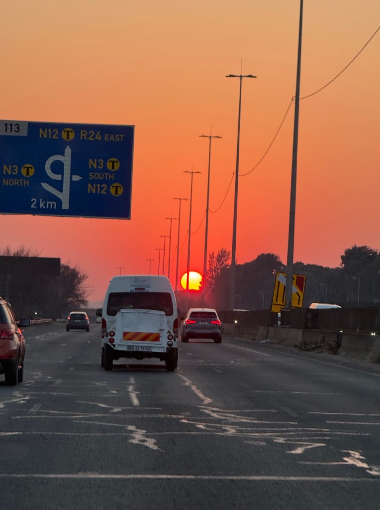 A more distant picture of the dull golden disc of the sun rising into a red, smoke filled sky. you can see the traffic on the road. 